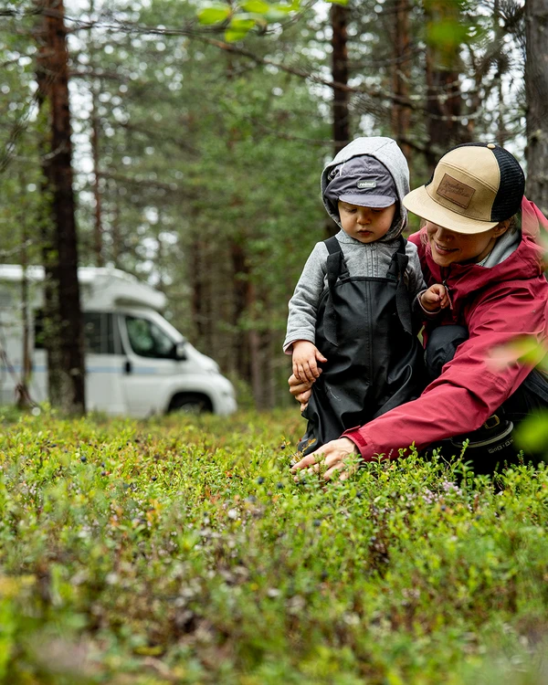Un adulto y un niño agachados en la hierba de un bosque, con una autocaravana detrás.