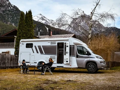 Autocaravana aparcada delante de una casa al pie de montañas nevadas, con dos personas en sillas de camping