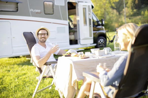 Pareja comiendo en una mesa puesta sobre la hierba delante de una autocaravana