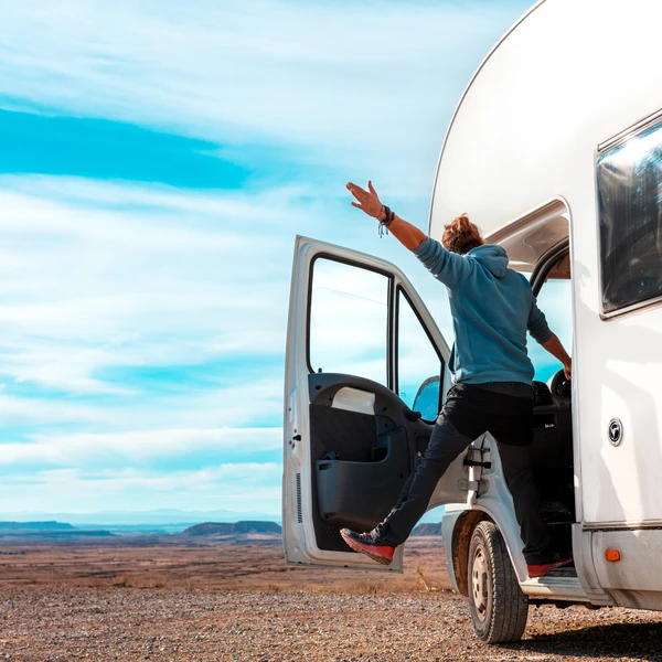 Viajero asomado por la puerta abierta de una autocaravana con un brazo en alto, en un paisaje desértico