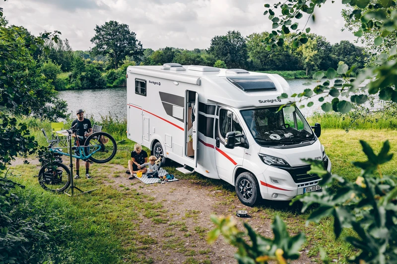 Familia junto a una autocaravana a la orilla de un río, con una bici en un soporte de taller y niños sobre una manta de pícnic