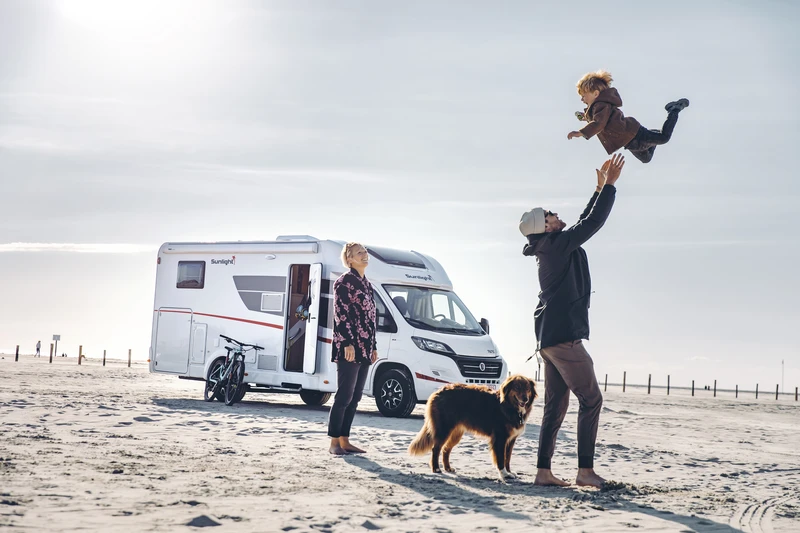 Padre lanzando a su hijo por los aires en una playa junto a una autocaravana, con la madre y un perro