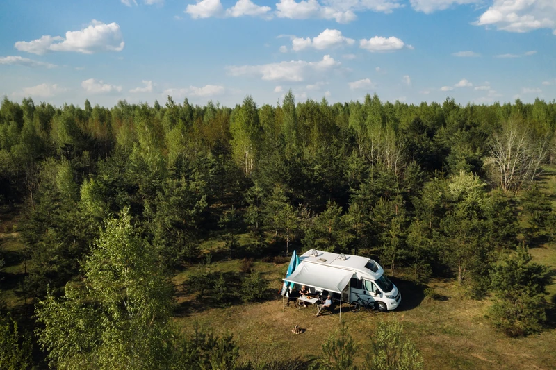 Vista aérea de una autocaravana con el toldo desplegado en un claro del bosque, con gente sentada a una mesa