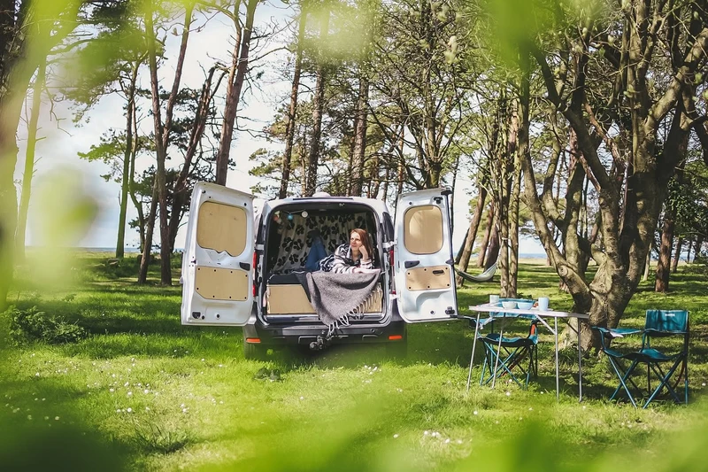 Viajera descansando bajo una manta en la parte trasera de una camper con las puertas abiertas, entre árboles, con mesa y sillas de camping