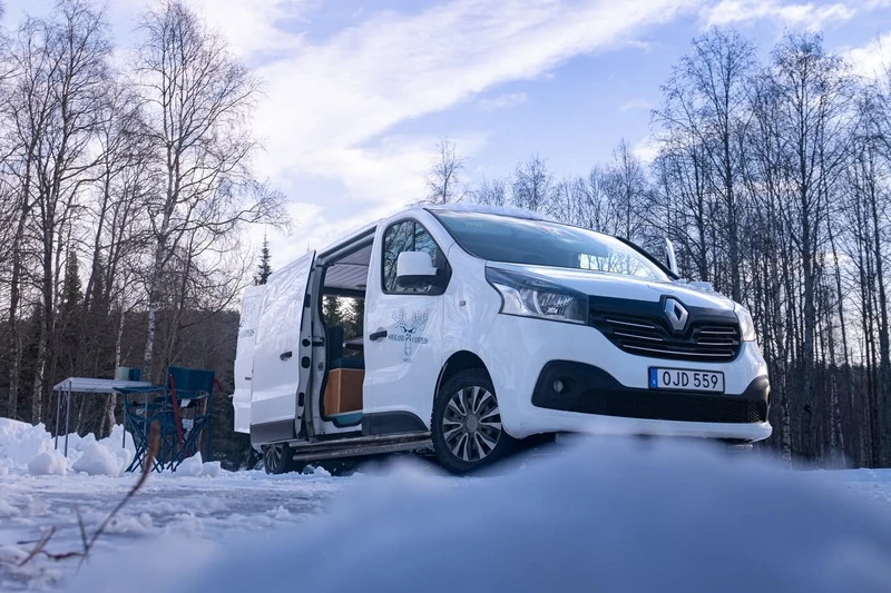 Camper con la puerta lateral abierta en un bosque de abedules nevado, con mesa y sillas de camping al lado