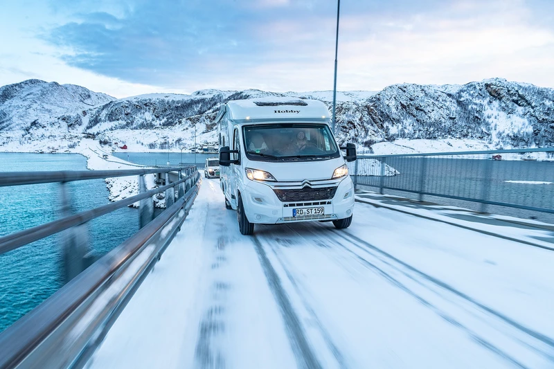 Autocaravana cruzando un puente nevado sobre el mar, con montañas nevadas al fondo