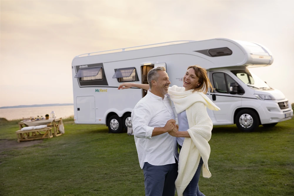 Pareja bailando en la hierba delante de una autocaravana junto al agua al atardecer