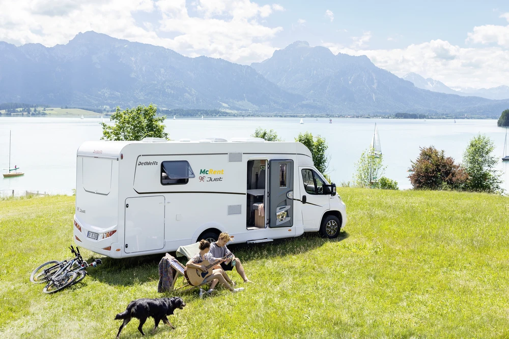 Pareja tocando la guitarra junto a una autocaravana en un prado a orillas de un lago, con montañas al fondo, bicicletas y un perro