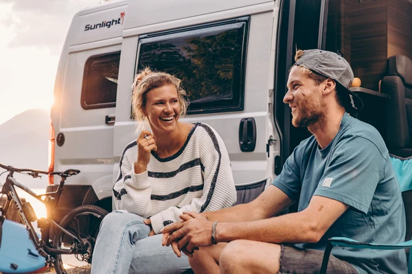 Pareja sonriente sentada delante de su camper, con una bicicleta al lado