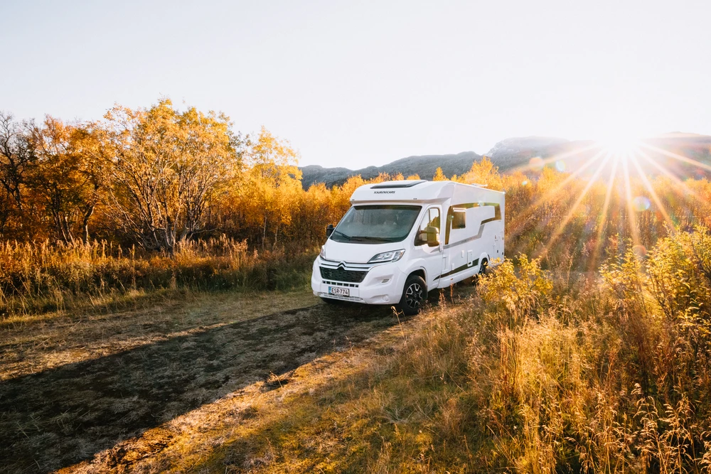Autocaravana en un camino de tierra entre matorrales dorados de otoño, con el sol bajo detrás