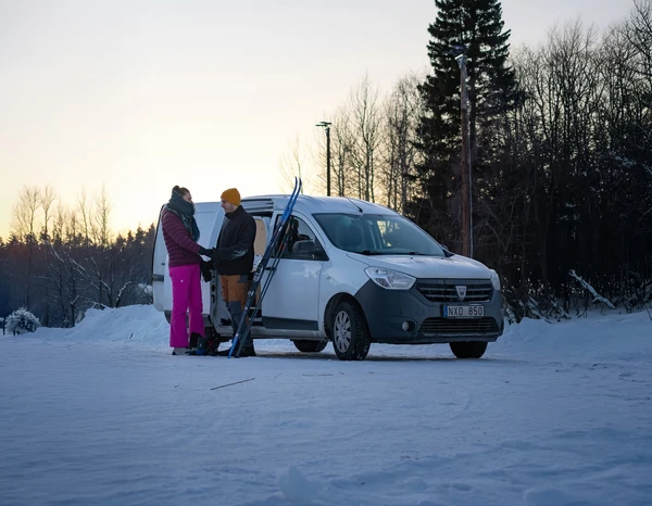Dos personas con esquís junto a una camper en una carretera nevada