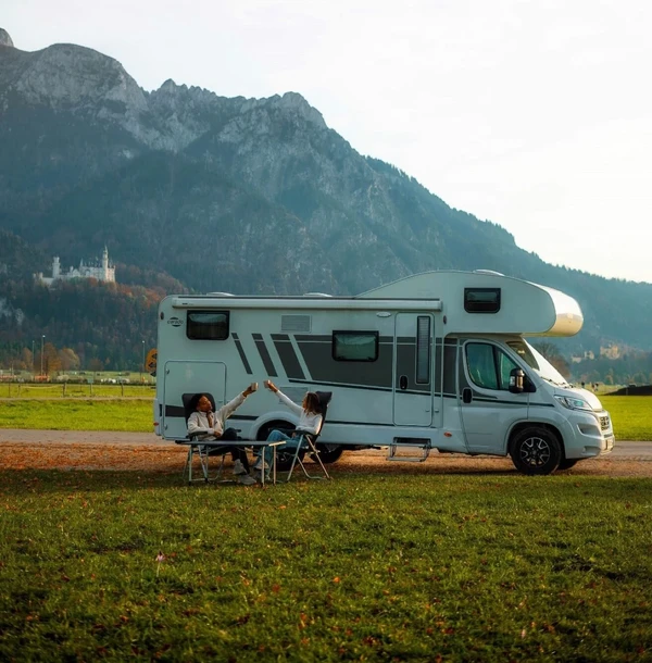 Autocaravana aparcada en un prado al pie de las montañas, con dos personas en una mesa de camping