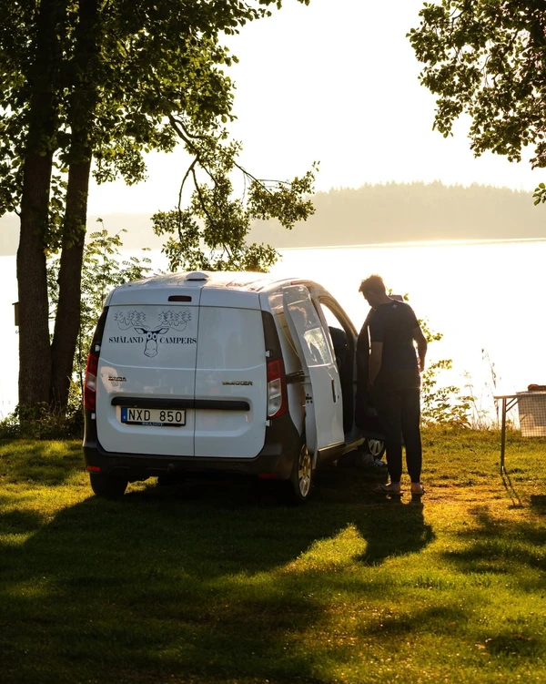 Camper bajo los árboles junto a un lago al atardecer, con una persona de pie al lado