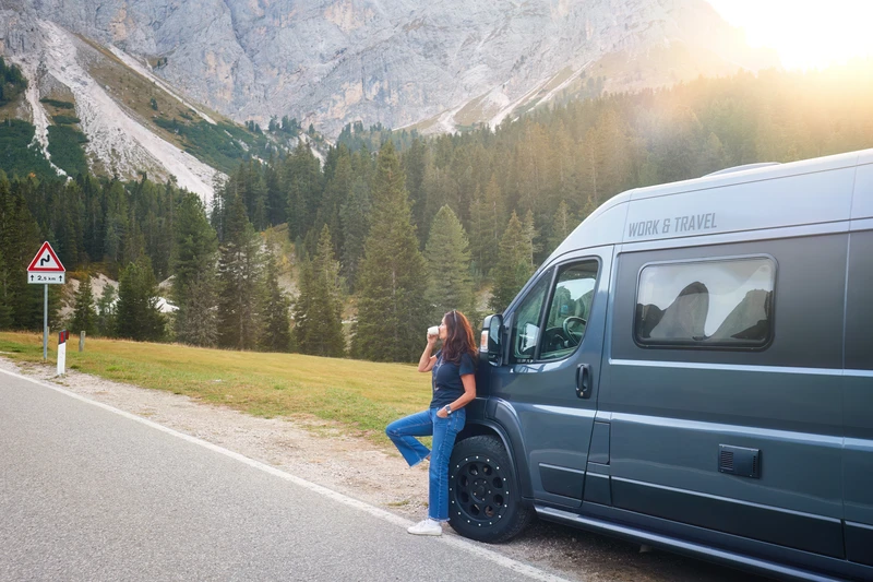 Mujer con una bebida caliente apoyada en una camper junto a una carretera de montaña