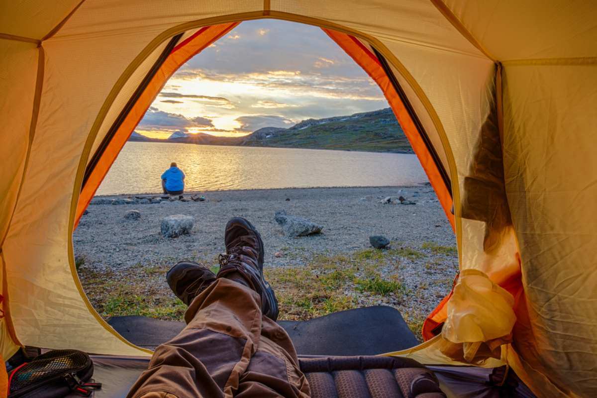 View from inside a tent looking out at a quiet lakeshore at sunset, with hiking boots in the foreground and a camper sitting by the water.