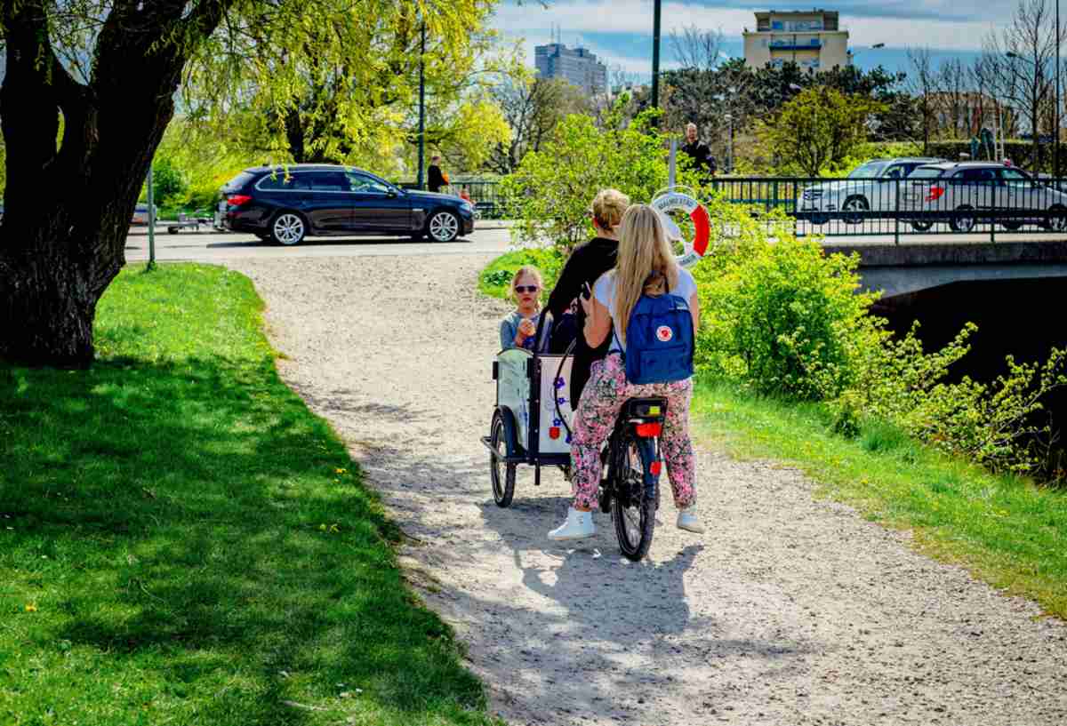 En familj bestående av en mamma och hennes två döttrar cyklar på en cykel i en stadspark i Malmö.
