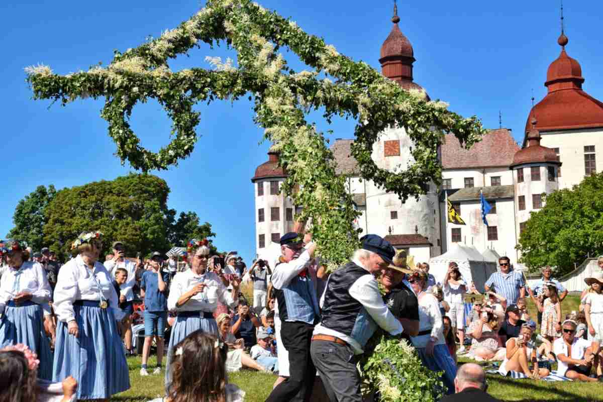 En grupp människor klädda i typiska svenska kläder reser ett blomsterkors för att markera sommarens ankomst.