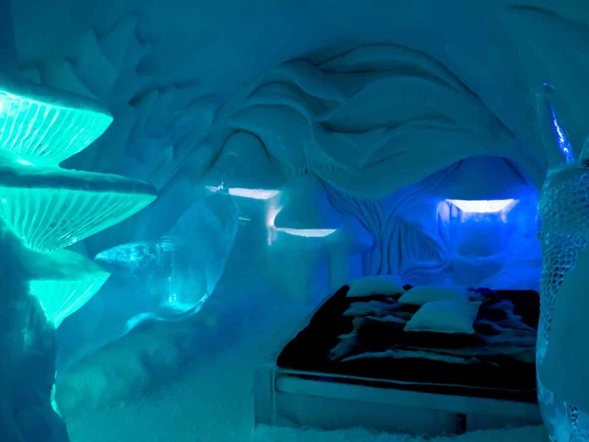View of the interior of one of the bedrooms at Sweden's famous Icehotel, completely covered in ice and illuminated by lights that seem to recreate the northern lights of the area, with a black bed.