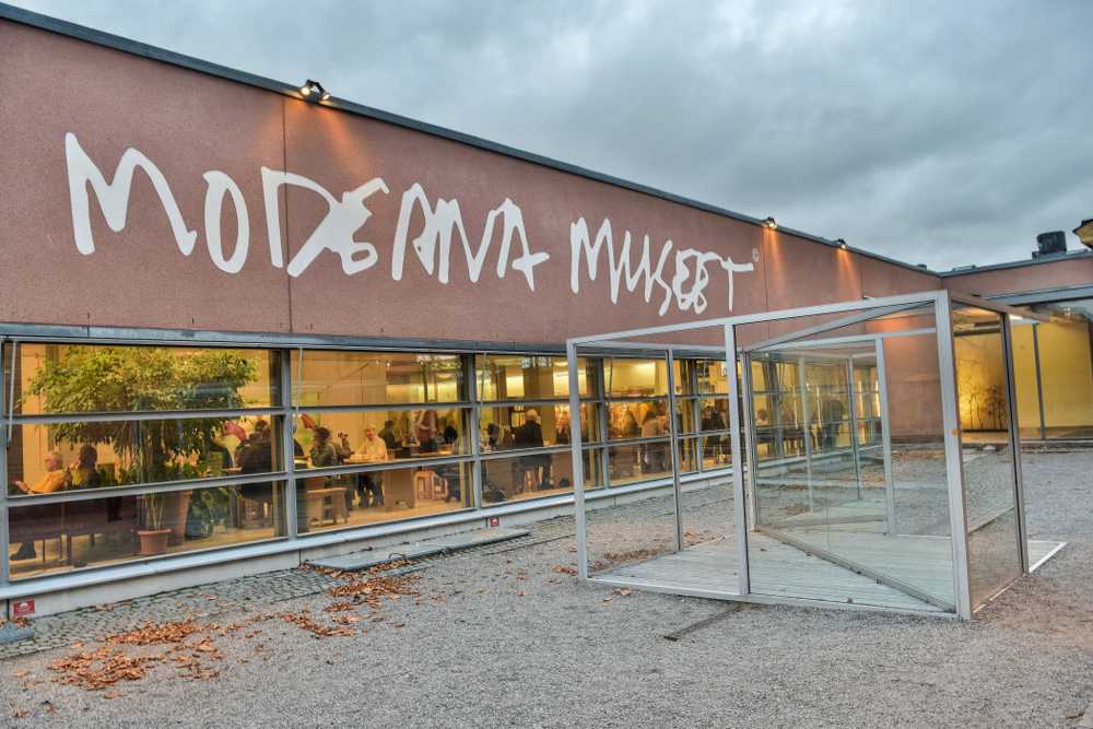 Overview of the façade of the Moderna Museet in Stockholm with several people sitting in what appears to be a dining room lit by warm light from inside through large windows.