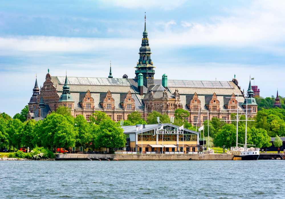 General view of the building that houses the Nordiska Museet in Stockholm from across a large body of water surrounded by trees, with a few boats moored in front of it.