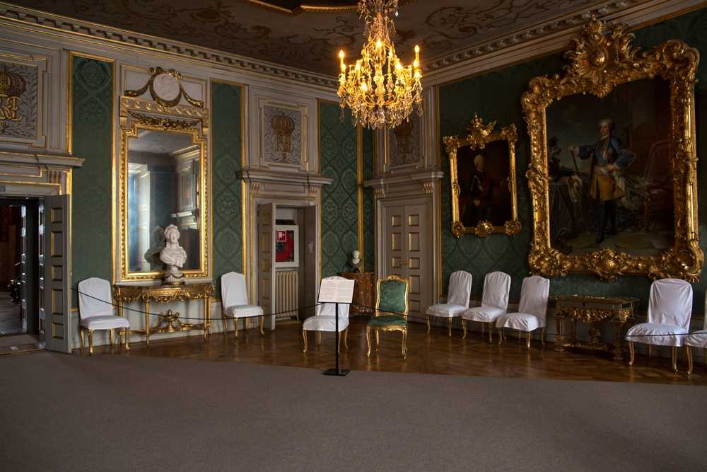 Detailed view of one of the rooms in the Royal Palace in Stockholm, featuring several upholstered chairs, enormous paintings with gilded frames, and a mirror in front of which stands a white sculpture.
