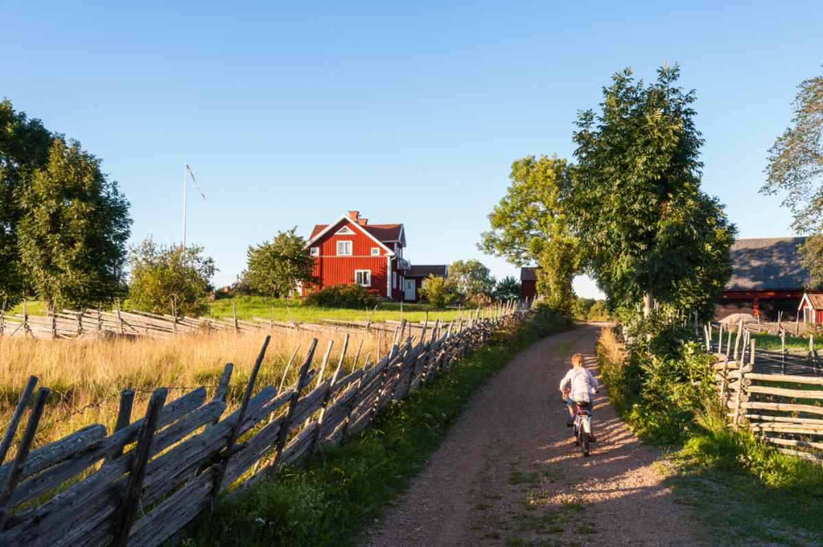 Ein Junge läuft auf einem Feldweg neben einem typisch schwedischen Bauernhaus, das an einem sonnigen Tag vollständig eingezäunt ist.