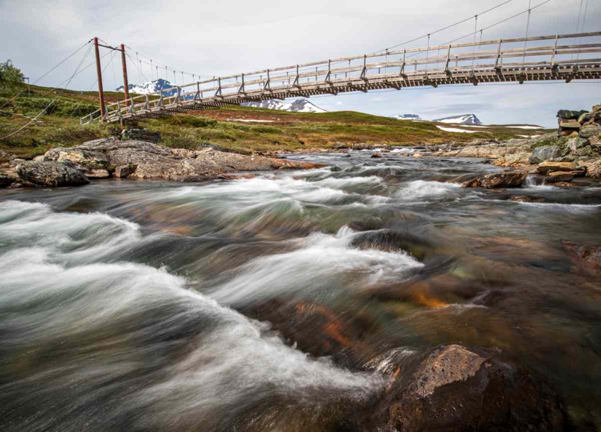 General view of a wooden bridge over a river with very strong water currents in a natural setting with snow-capped mountains in the background.