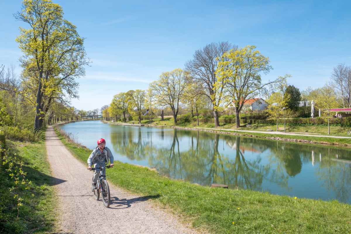 Person som cyklar längs Göta kanals dragväg en solig dag, med stilla vatten som speglar gröna träd och blå himmel.
