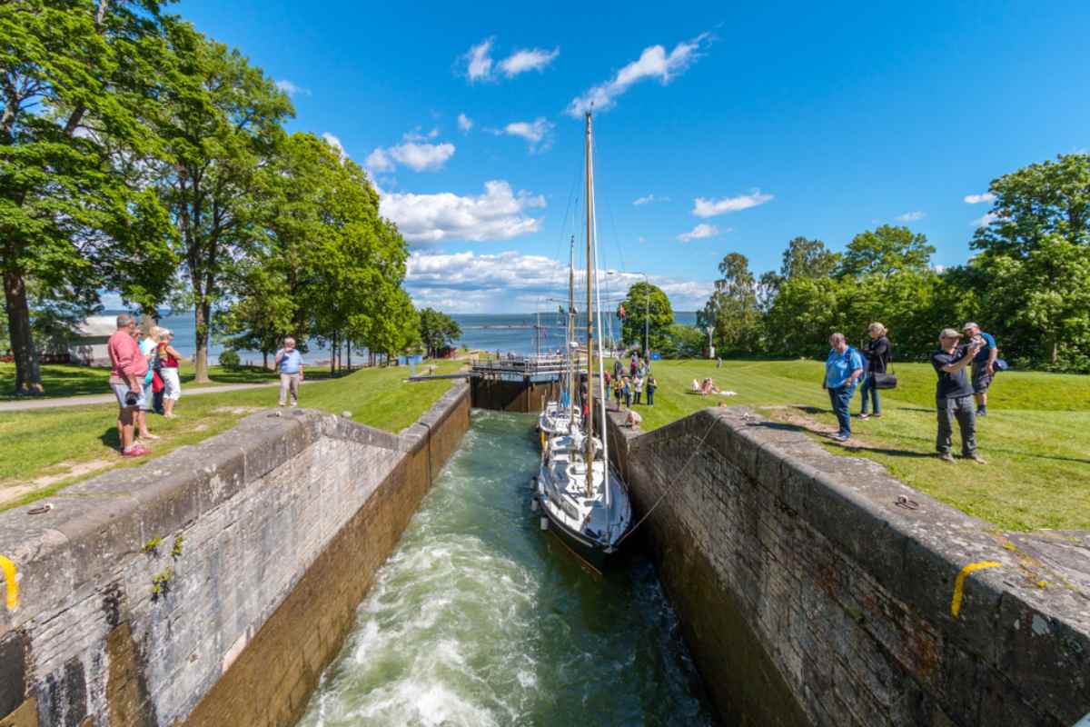Segelbåt som passerar en sluss i Göta kanal medan besökare tittar från gräsbankar och stenmur en solig dag.