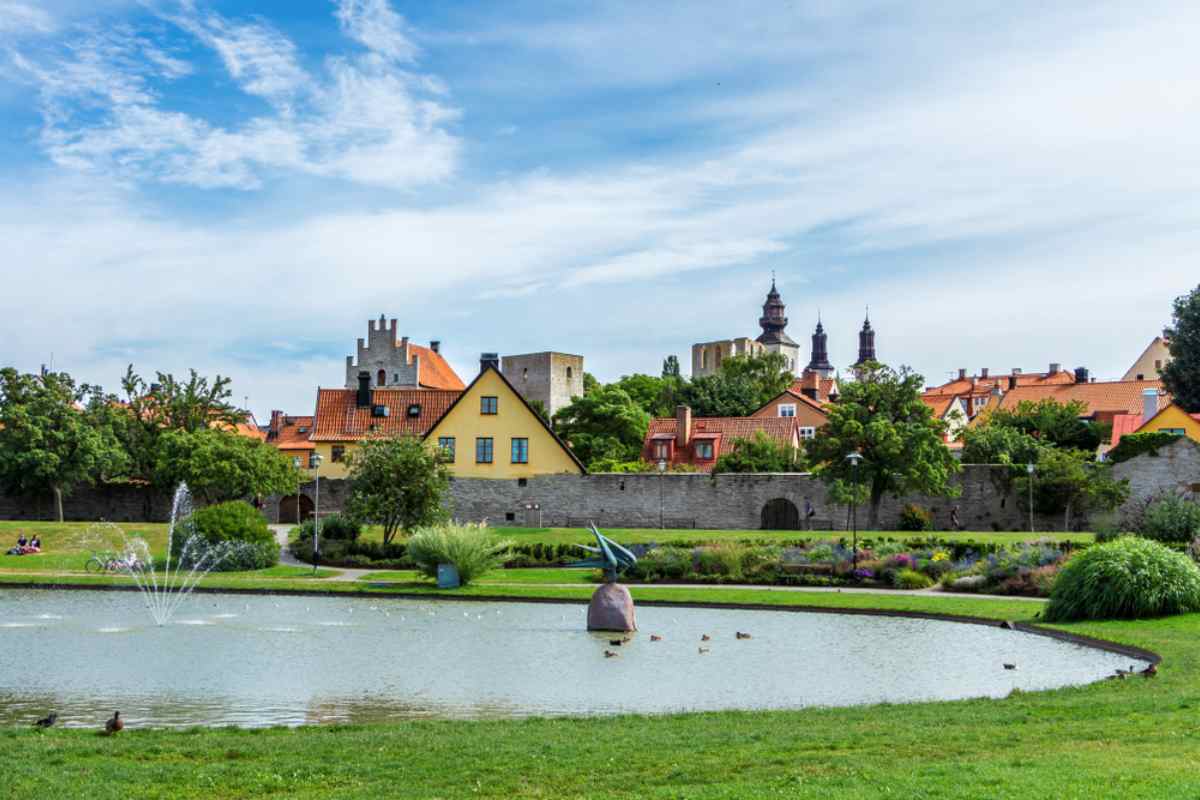 Malersiche Ansicht des Almedalen-Parks in Visby, Schweden, mit bunten Häusern, mittelalterlichen Mauern und einem Teich mit Fontänen.