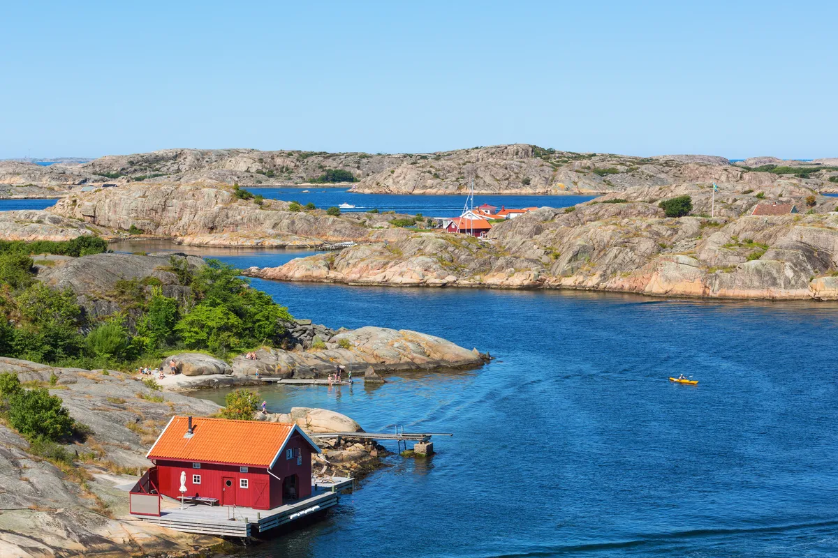 Rött båthus på den steniga kusten i den svenska skärgården med lugnt blått vatten och utspridda stugor.