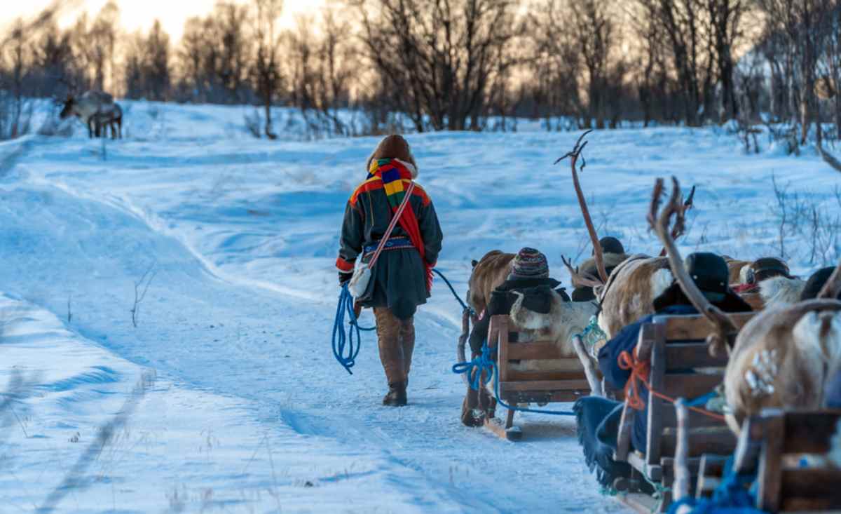 En samisk person drar en släde med flera renar i ett helt snötäckt svenskt landskap.
