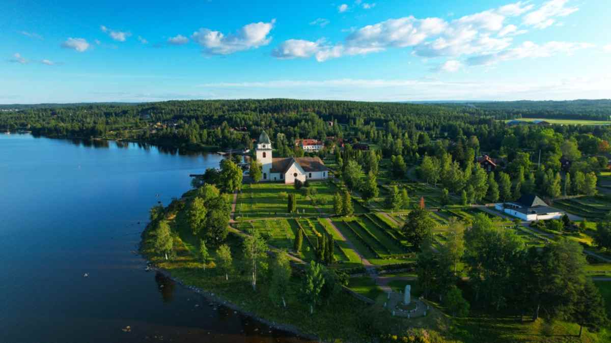 Aerial of a white country church and patterned gardens beside a still lake, surrounded by forest in Dalarna, Sweden.
