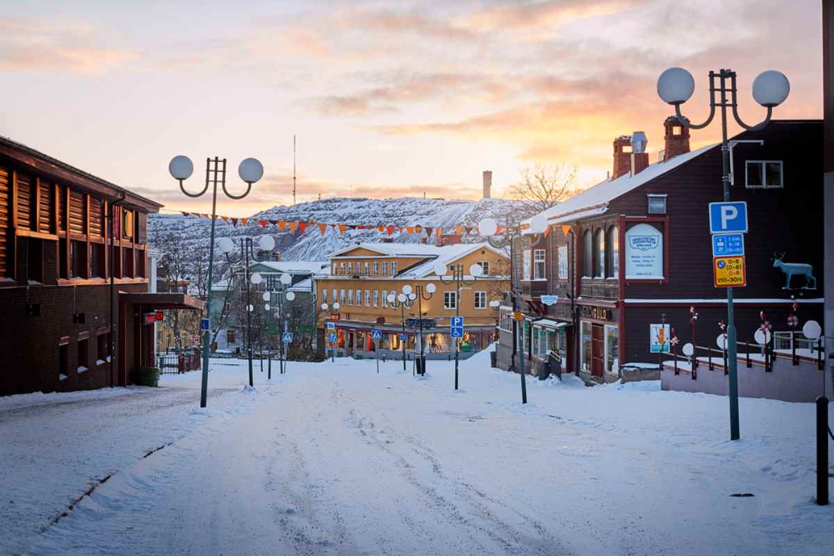 Snowy main street in Kiruna at sunset, lined with wooden buildings and streetlamps, with hills in the background.