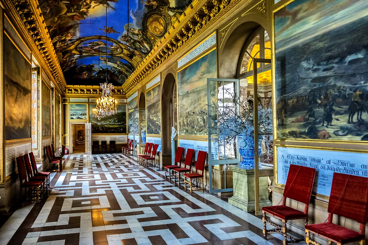 Interior of Drottningholm Palace showcasing a grand hall with ornate ceiling paintings, chandeliers, and luxurious red chairs