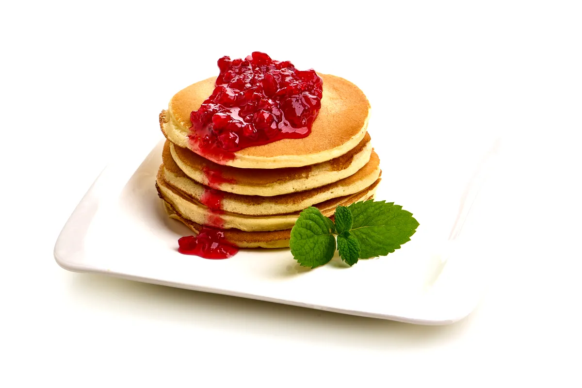 A stack of fluffy pancakes topped with lingonberry sauce, garnished with a fresh mint leaf on a white square plate.