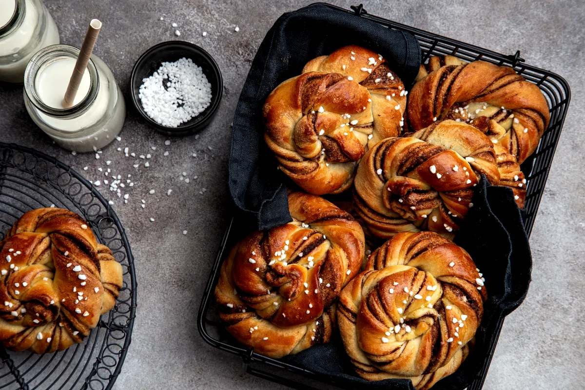 En korg med nybakade svenska kanelbullar beströdda med pärlsocker, tillsammans med flaskor med mjölk och en skål med socker på en mörk bänkskiva