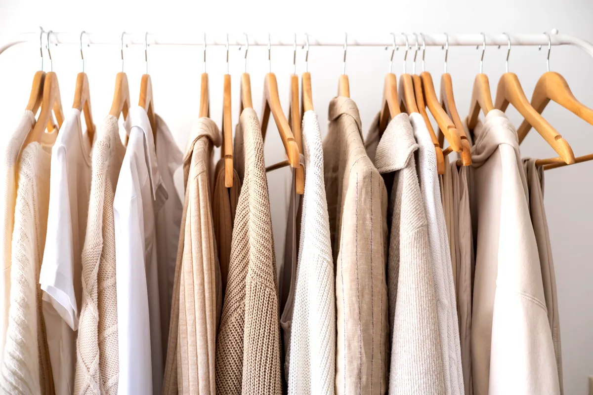 Ett urval av klädesplagg i neutrala toner, inklusive beige, vitt och krämfärgad, snyggt upphängda på trägalgar på ett vitt metallställ. Kläderna varierar i struktur, med vissa delar gjorda av stickat, linne och andra mjuka tyger , vilket skapar en sammanhängande och minimalistisk estetik. Den övergripande paletten är varm och lugnande, och återspeglar en enkel, diskret stil. Scenen är väl upplyst och framhäver plaggens strukturer och färger, som är arrangerade för att skapa en balanserad och harmonisk visning. 