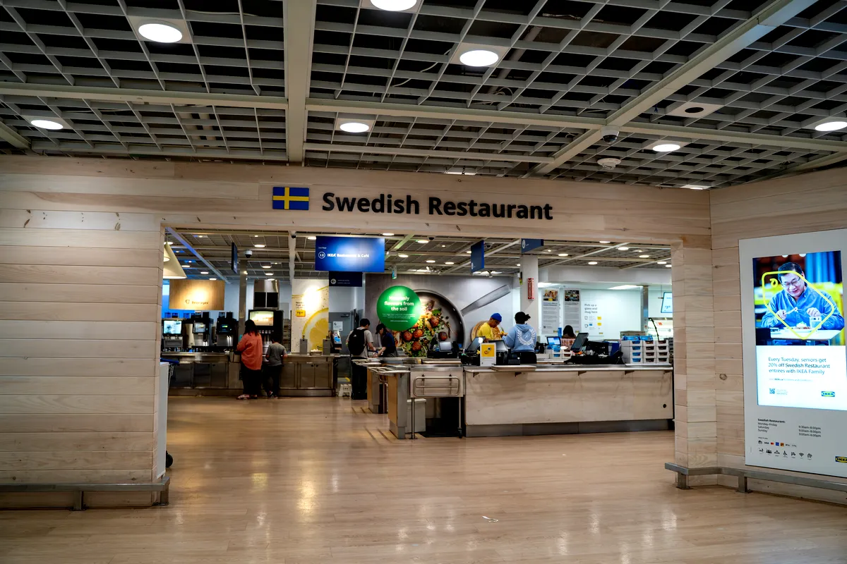Entré till en svensk restaurang inne i ett IKEA varuhus, kännetecknad av minimalistisk design och ljusa trätoner. På skylten ovanför entrén står det 