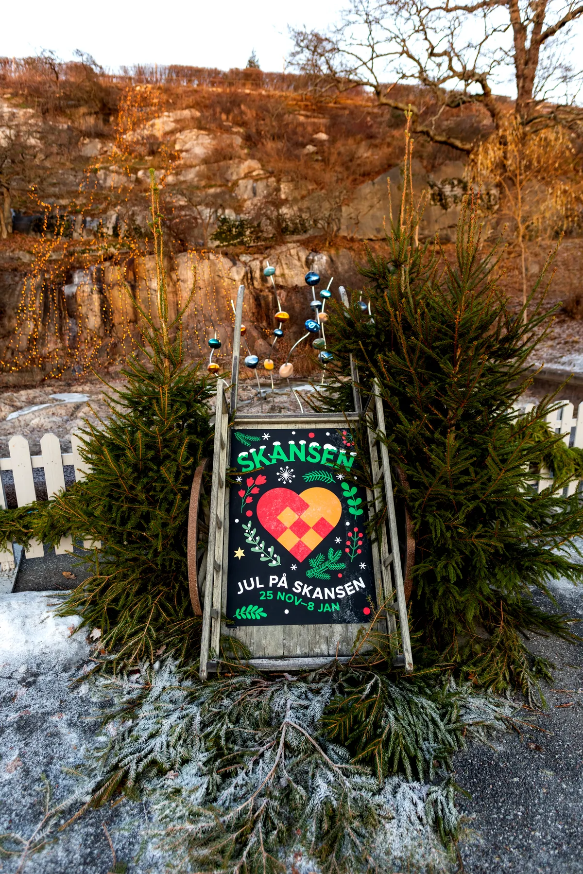 A festive Christmas-themed sign at Skansen, set up in a natural outdoor setting with pine trees and holiday decorations. The sign features a colorful woven heart design with the words “Skansen” at the top, and the text “Christmas at Skansen, November 25 – January 8” at the bottom. The display is surrounded by winter greenery and a frosted landscape, suggesting a cozy, seasonal atmosphere.