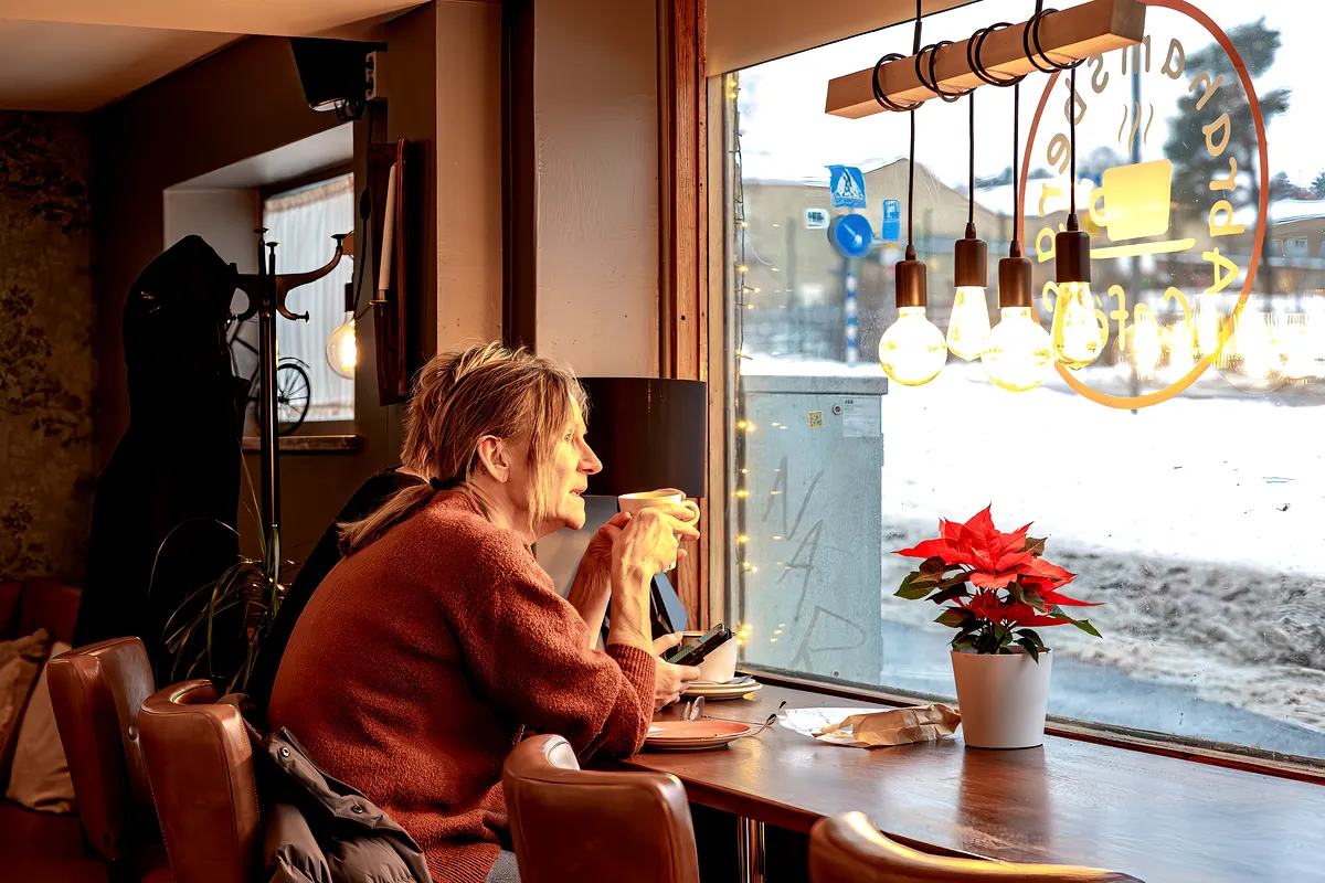 A woman sitting by the window of a cozy café in Stockholm, Sweden. She holds a warm cup of coffee in her hands while gazing outside, with a relaxed expression on her face. The café has a warm, inviting atmosphere, with hanging lightbulbs and a poinsettia plant on the table. Snow is visible outside the window, adding to the cozy ambiance of the scene.