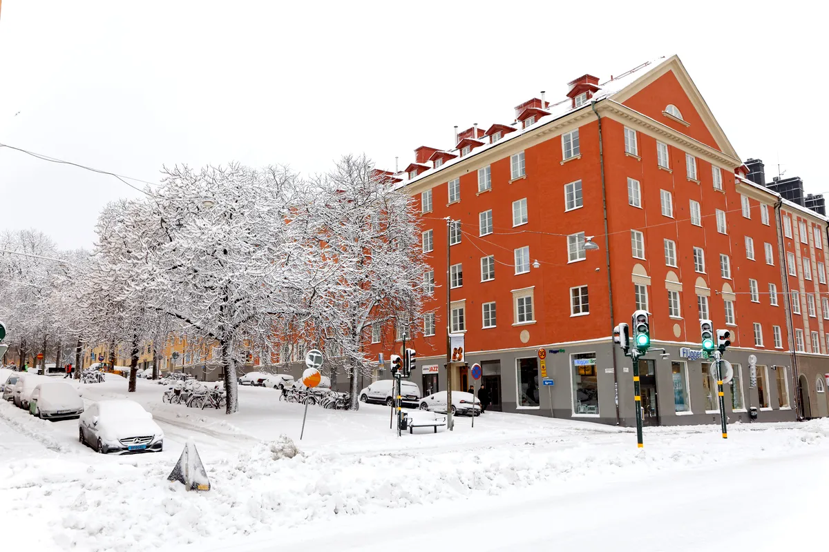 A street in Stockholm covered in a thick layer of snow during winter. The scene shows snow-covered cars, trees, and buildings, with a large, red apartment block as the focal point. The trees’ branches are heavily laden with snow, creating a peaceful, wintry atmosphere. Pedestrians are walking along the snowy sidewalks, and traffic lights are glowing, signaling a functioning city despite the weather.