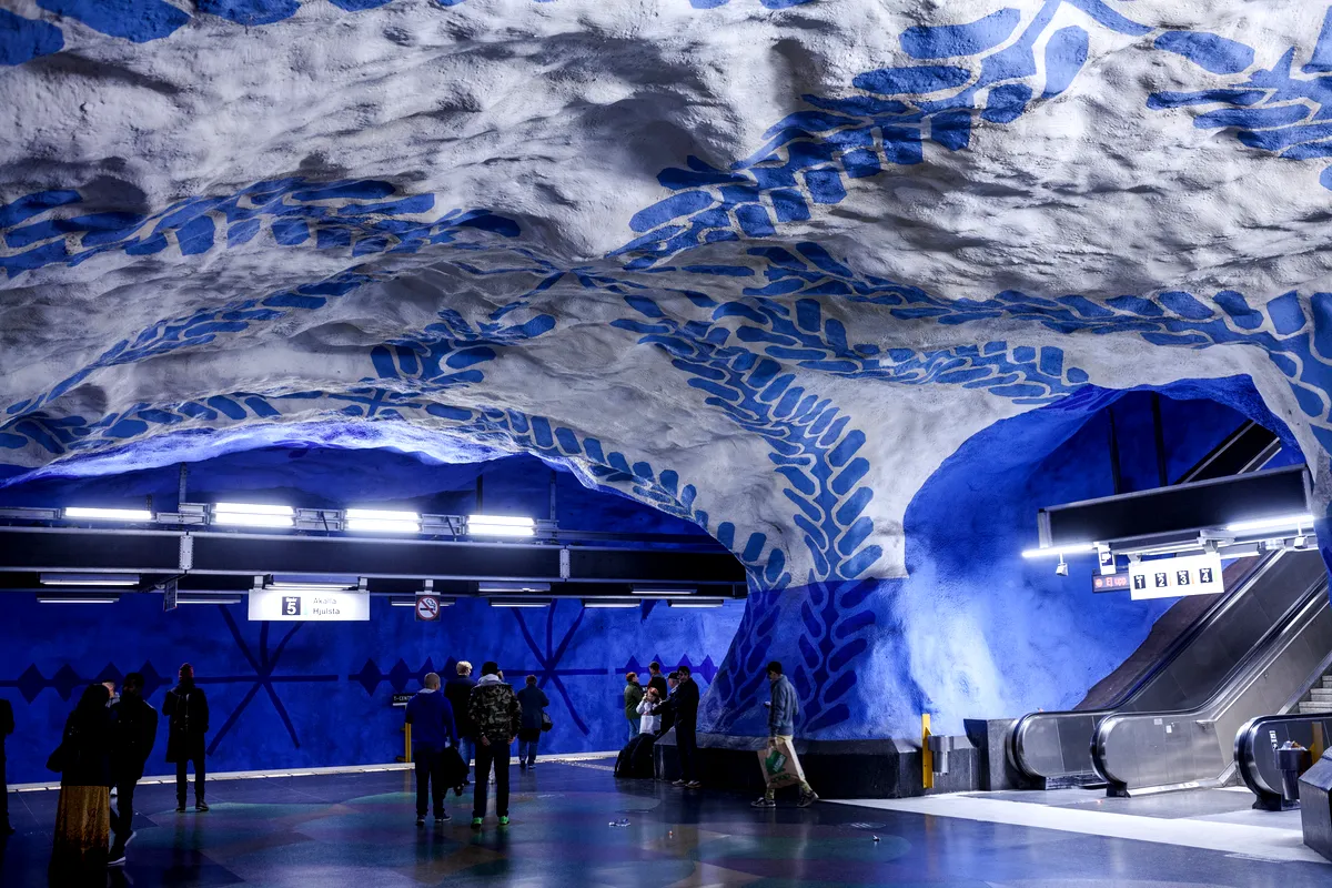 Ett foto av interiören av en tunnelbanestation i Stockholm på Blå linjen, med imponerande grottliknande väggar målade i vitt och blått. Väggarna har djärva mönster som liknar löv eller organiska former, vilket skapar en unik och konstnärlig miljö. Pendlare är står och går nära en rulltrappa, och stationens starka belysning lägger till kontrast till de levande färgerna.