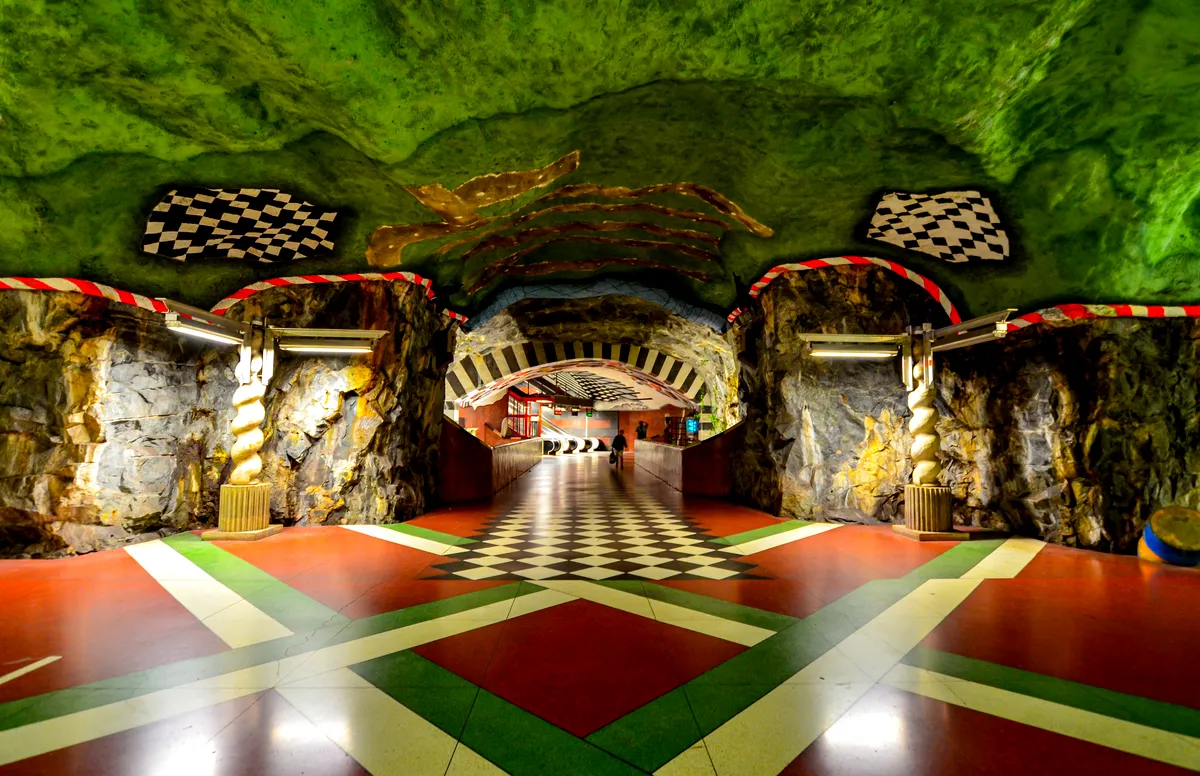 Ett fotografi av Kungsträdgårdens station i Stockholms tunnelbanesystem. Väggarna och taket är designade för att likna en grotta, målade i en slående grön färg. Golvet är ett färgglatt geometriskt mönster i rött, grönt, svart och vitt, vilket skapar En levande kontrast flankeras av stenpelare med vridna mönster, som leder in i en tunnel med svarta och vita rutiga mönster i taket. width=