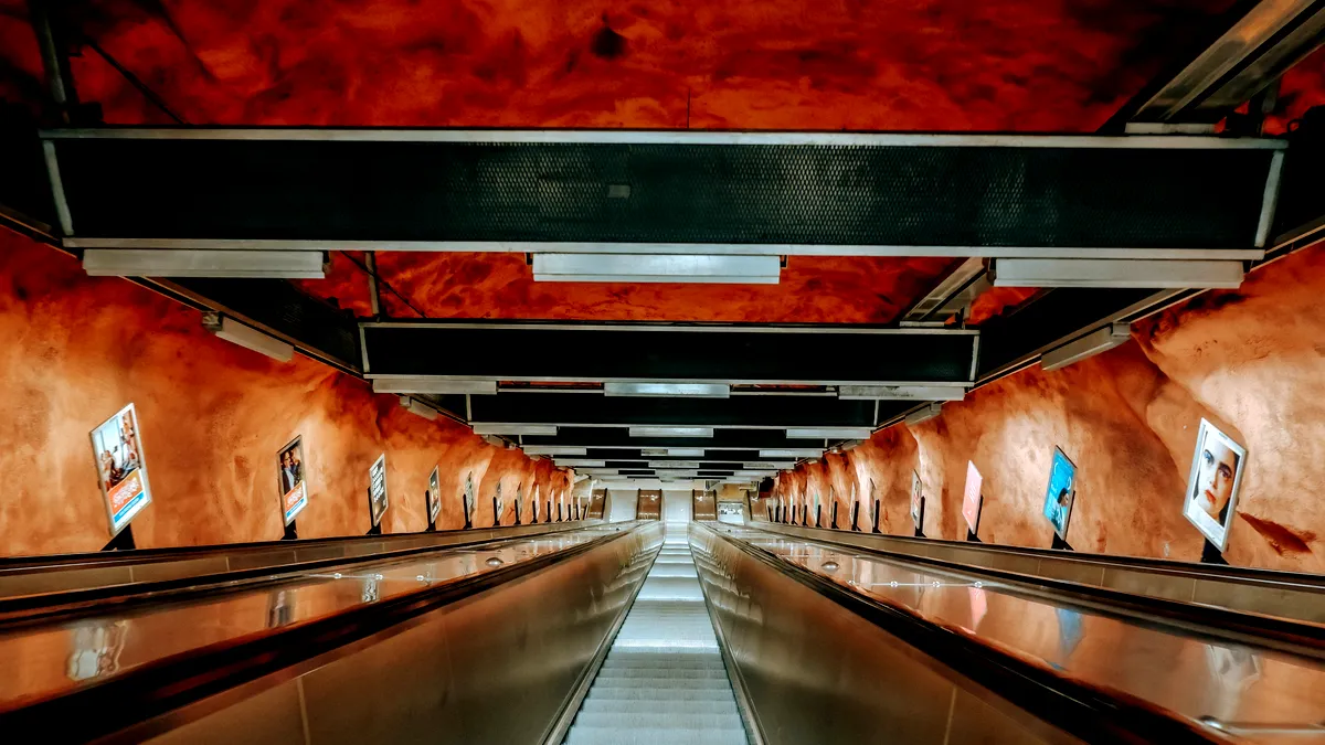 En lång, dramatisk rulltrappa vid Rådhusets tunnelbanestation i Stockholm, Sverige. Stationens väggar och tak är målade i djupa nyanser av rödbrunt, som liknar en grotta. Metallbalkar och belysningsarmaturer hänger ovanför, vilket bidrar till Industriell och grottlik atmosfär kantas av reklam på båda sidor av rulltrappan, som verkar sträcka sig långt bort. width=