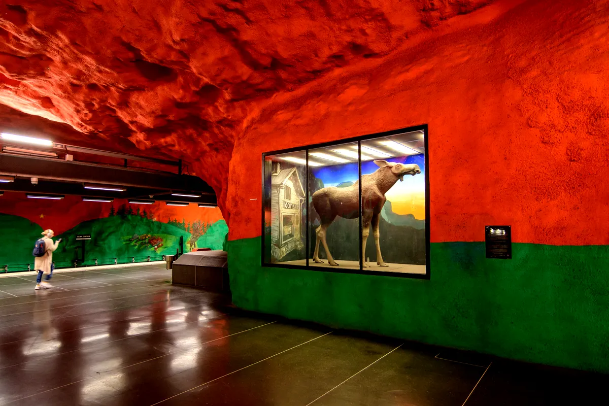 En färgstark underjordisk konstinstallation vid Solna Centrums tunnelbanestation i Stockholm. Stationens väggar och tak är målade i levande rött och grönt, som liknar en grotta. En glasmonter till höger innehåller en modell av en älg i naturlig storlek, mot bakgrund av ett lantligt hus och en skog, med en stjärnklar natthimmel, en person ses gå i bakgrunden till vänster och ta in de livfulla väggmålningarna som skildrar naturscener och traditionella svenska landskap. width=