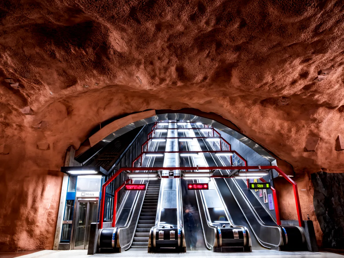 Ett foto av tunnelbanestationen Stockholm Stadion, med rulltrappor som leder uppåt genom ett grottliknande tak målat i en djupt rödbrun nyans. Väggarnas naturliga stenstruktur ger stationen en rå, organisk känsla. De moderna rulltrapporna , omgiven av röda metallräcken, kontrasterar skarpt med den naturalistiska miljön. Pendlare kan ses röra sig på rulltrapporna, och stark belysning lyser upp utrymmet.
