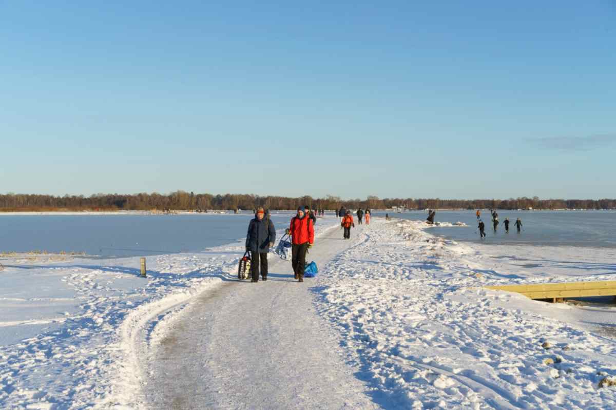 Eine Gruppe von Menschen, einige auf einem zugefrorenen See und andere auf einem verschneiten Weg, genießen einen sonnigen Tag beim Eislaufen am Stadtrand von Schweden.