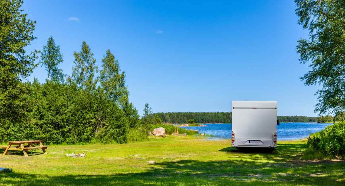 Ein weißer Camper ist in einer schwedischen Landschaft nahe einem See an einem wunderschönen, sonnigen Tag geparkt, an dem die Vegetation in voller Pracht ist.