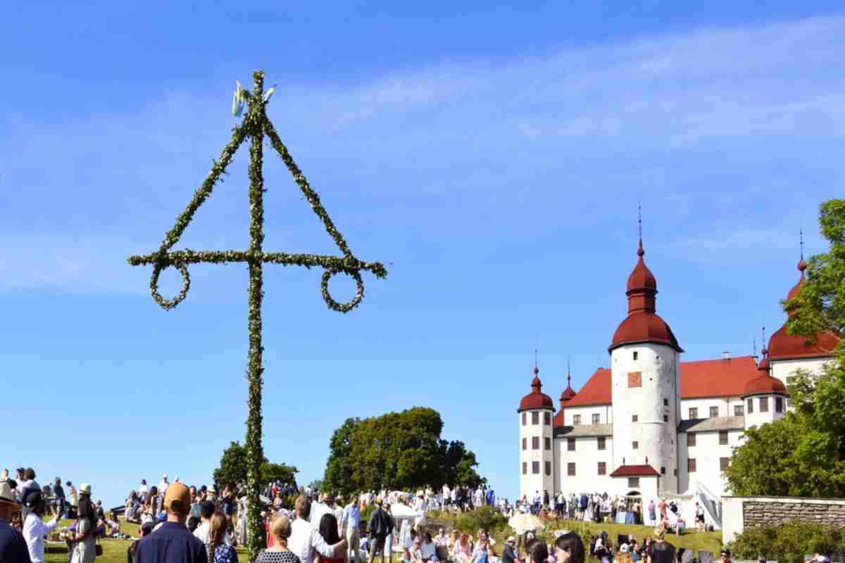 Typisches schwedisches Kreuz, das aus Blumen besteht und mitten auf einer grünen Wiese errichtet ist, wo eine Menschenmenge das Mittsommerfest feiert
